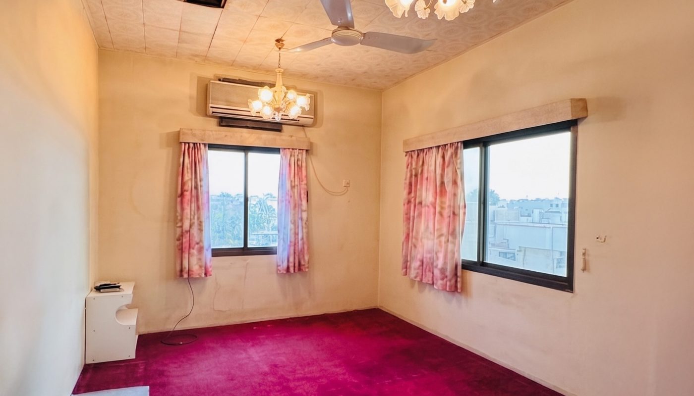 A sparsely furnished room with bright pink carpet, two windows with floral curtains, chandeliers, an AC unit, a ceiling fan, and a damaged ceiling panel.