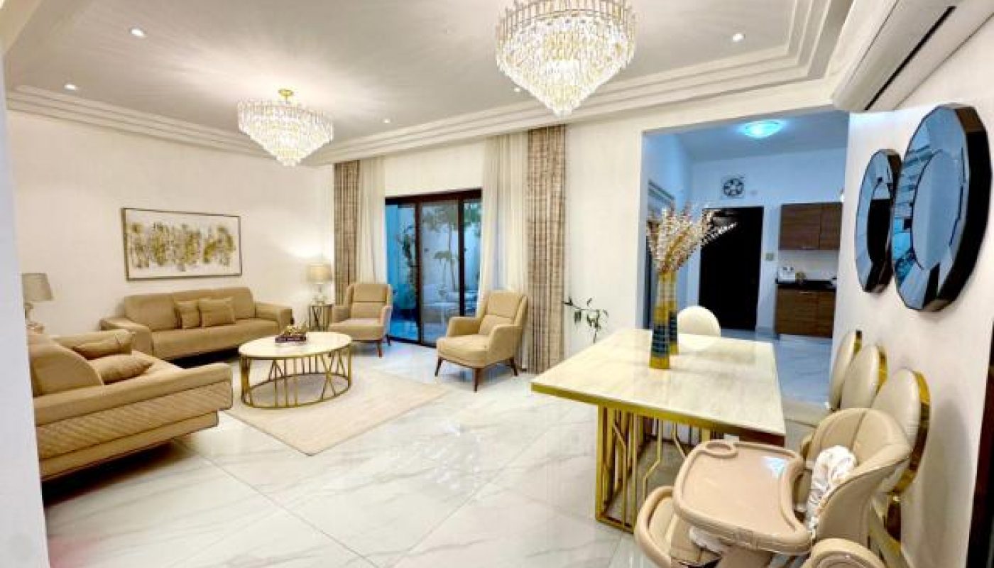 Spacious modern living and dining area with beige sofas, chandeliers, marble flooring, glass doors, and a baby high chair near a gold-accented dining table.