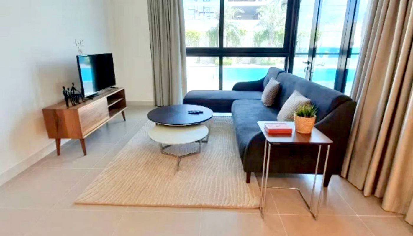 Modern living room with a grey sofa, round coffee table, TV on a wooden stand, side table with a plant, large windows, and beige tiled floor.