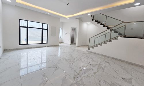 A spacious, bright room with white marble flooring, a staircase with glass railing, a large window, and recessed ceiling lighting in this luxurious villa for sale in Barbar.