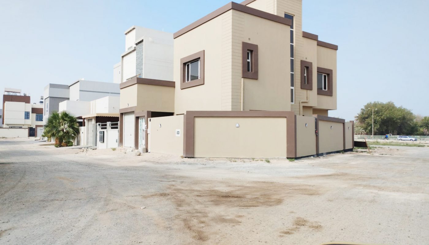Modern two-story beige house with brown trim situated on a corner lot, surrounded by a concrete wall, on a dry, unpaved street in a residential area.