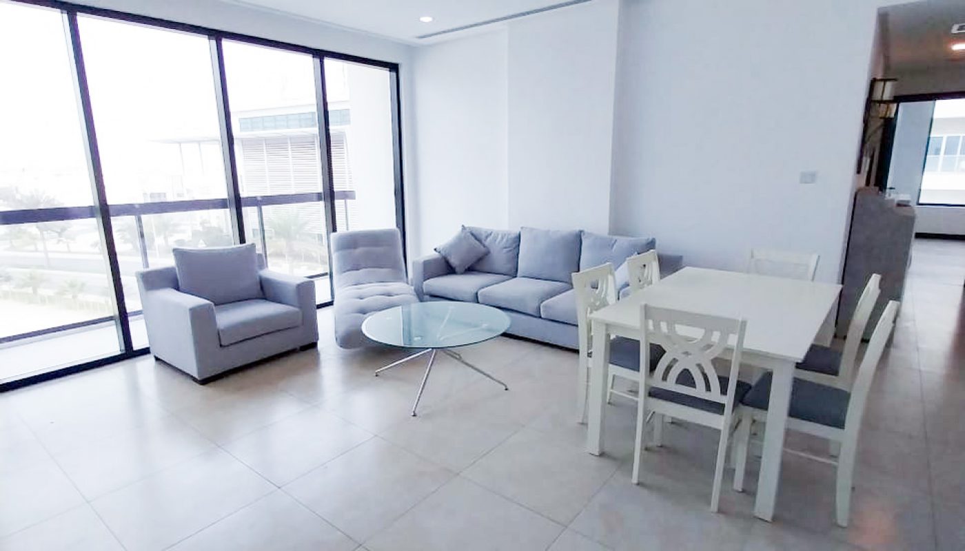 A modern, unfurnished living and dining area with large windows, a gray sofa set, glass coffee table, and white dining table with chairs on a tiled floor.