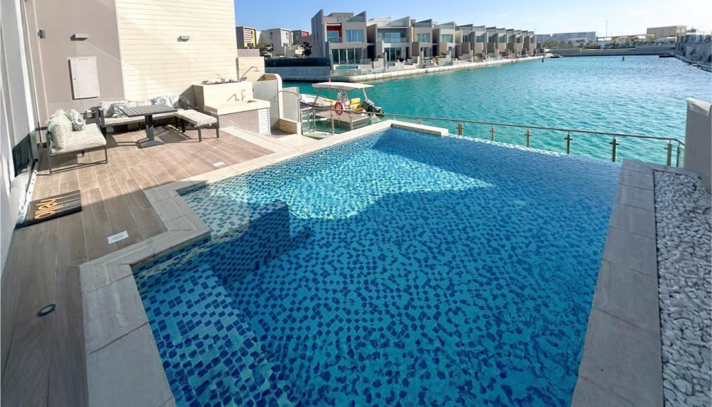 Infinity pool overlooking calm turquoise water, with lounge chairs and modern waterfront villas visible in the background under a clear blue sky.