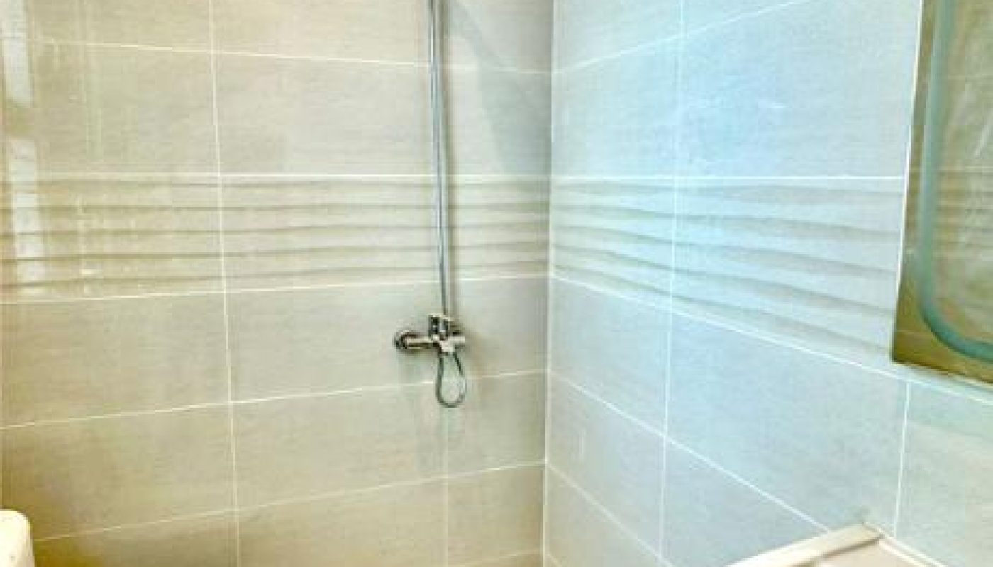 A bathroom with light-colored tiles, a wall-mounted shower, a toilet, and a sink. There is a small drain on the floor near the shower area.