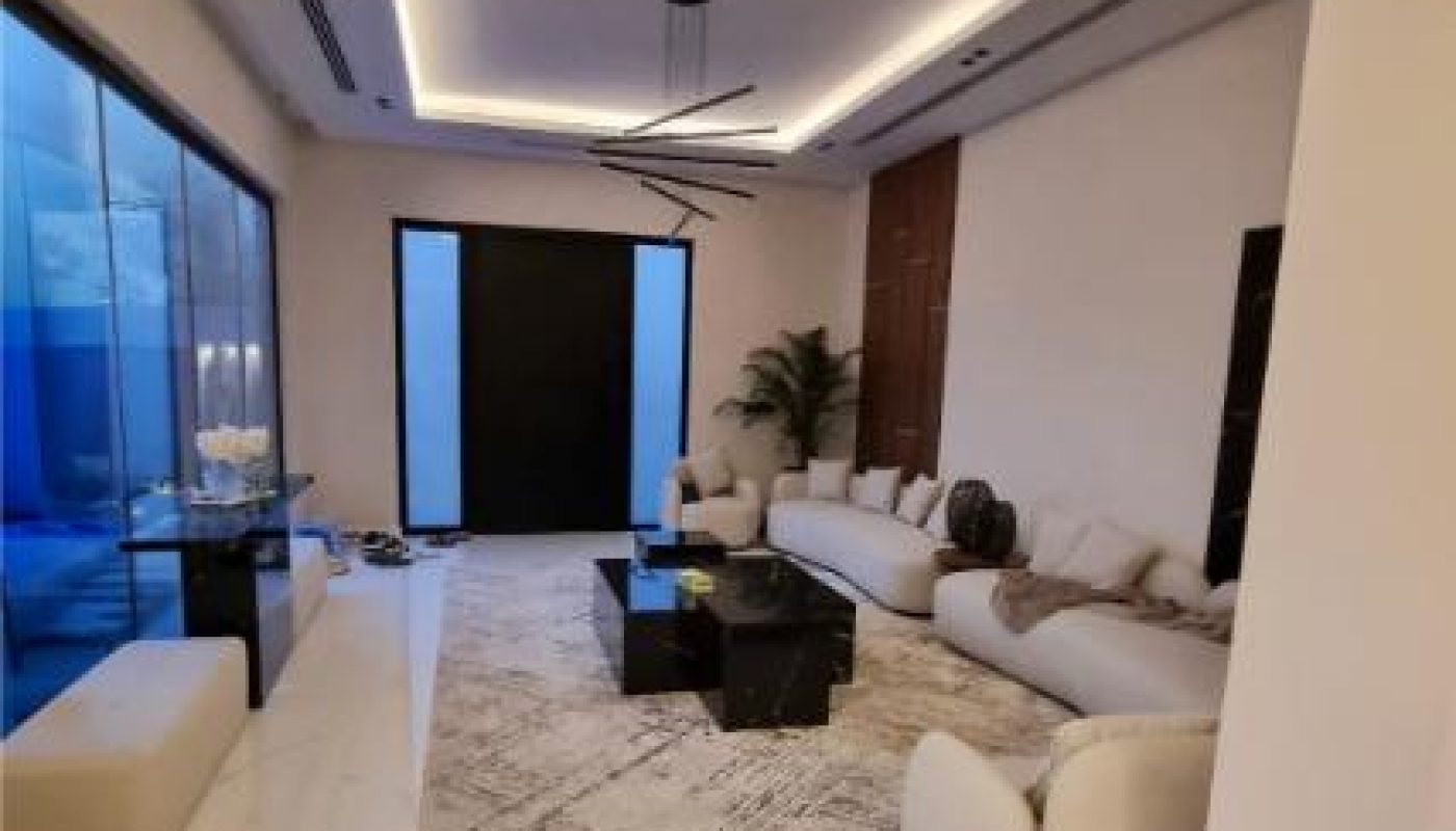 Modern living room with a long white sectional sofa, black coffee tables, a large area rug, minimalist decor, and floor-to-ceiling windows on one side.