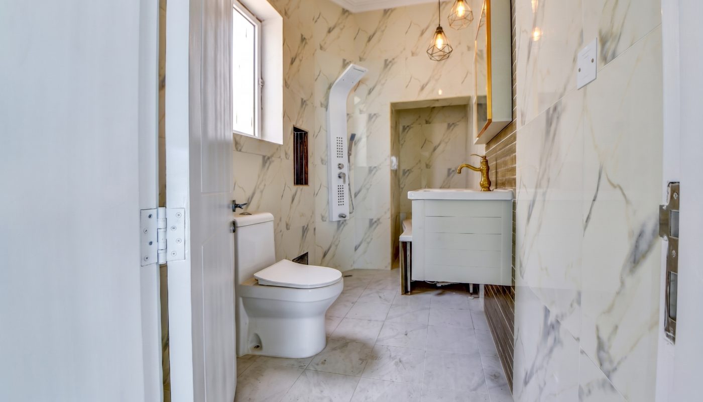 Modern bathroom with marble walls, a toilet, shower unit, white vanity with sink, gold faucet, wall mirror, and pendant lights.