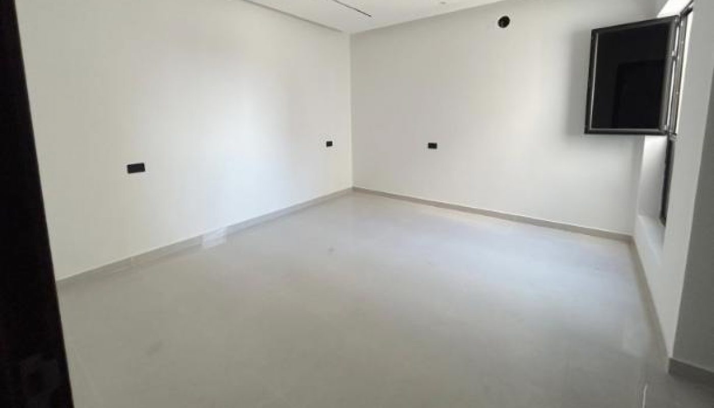 Empty room with white walls, gray floor, a ceiling with recessed lines, a single window on the right wall, and electrical outlets visible on the walls.