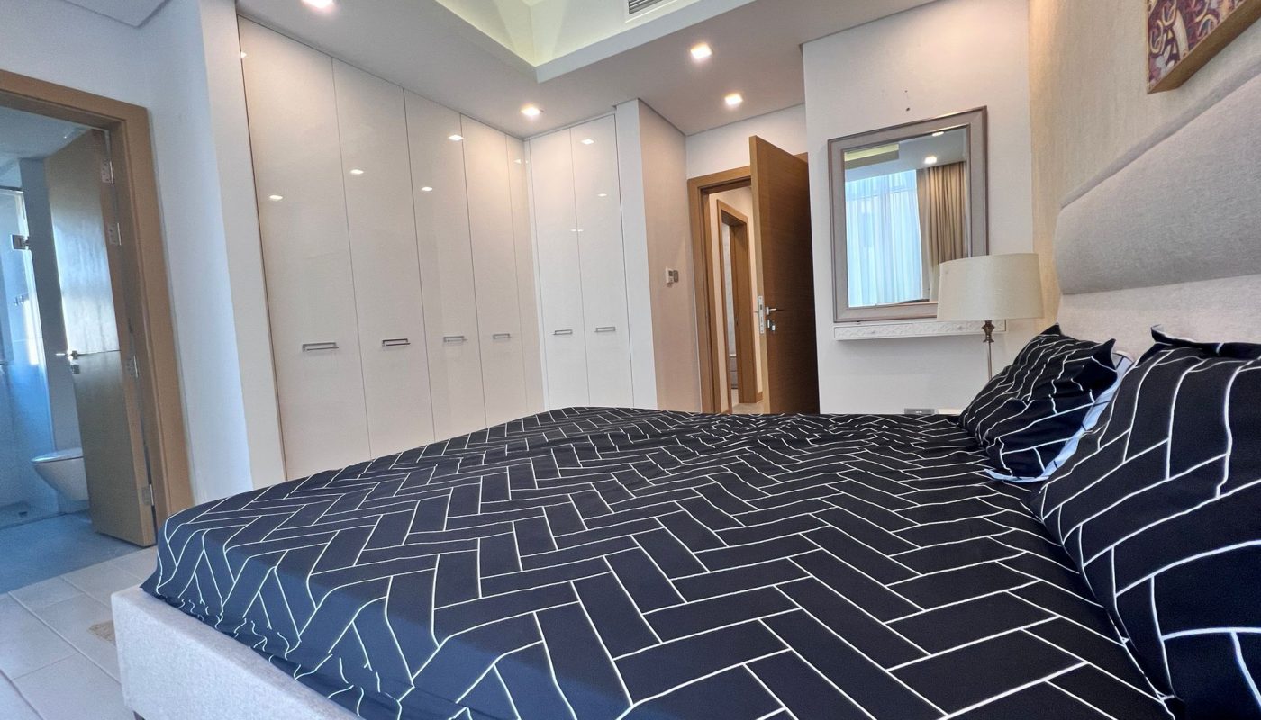 Modern bedroom with a geometric-patterned black bedspread, white wardrobes, a bedside lamp, and an open door revealing a bathroom.