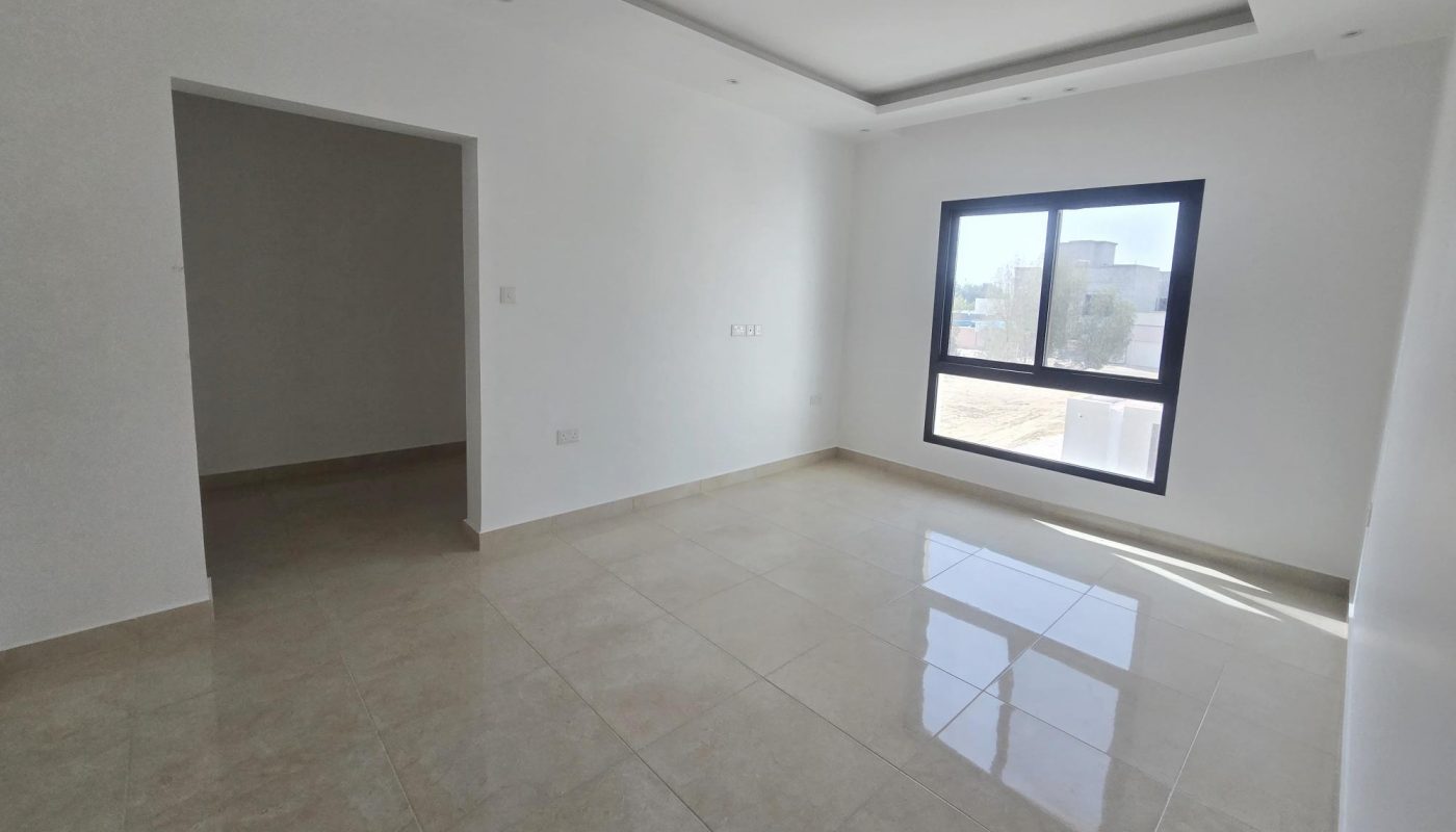 Empty, unfurnished room with white walls, tiled floor, a large window letting in sunlight, and an open doorway leading to another space.