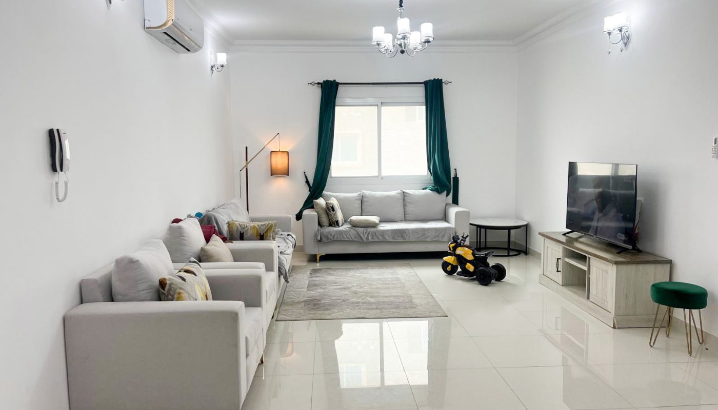 A modern living room with white walls, tile flooring, two sofas, a TV on a stand, a yellow toy bike, green curtains, and ceiling lights.