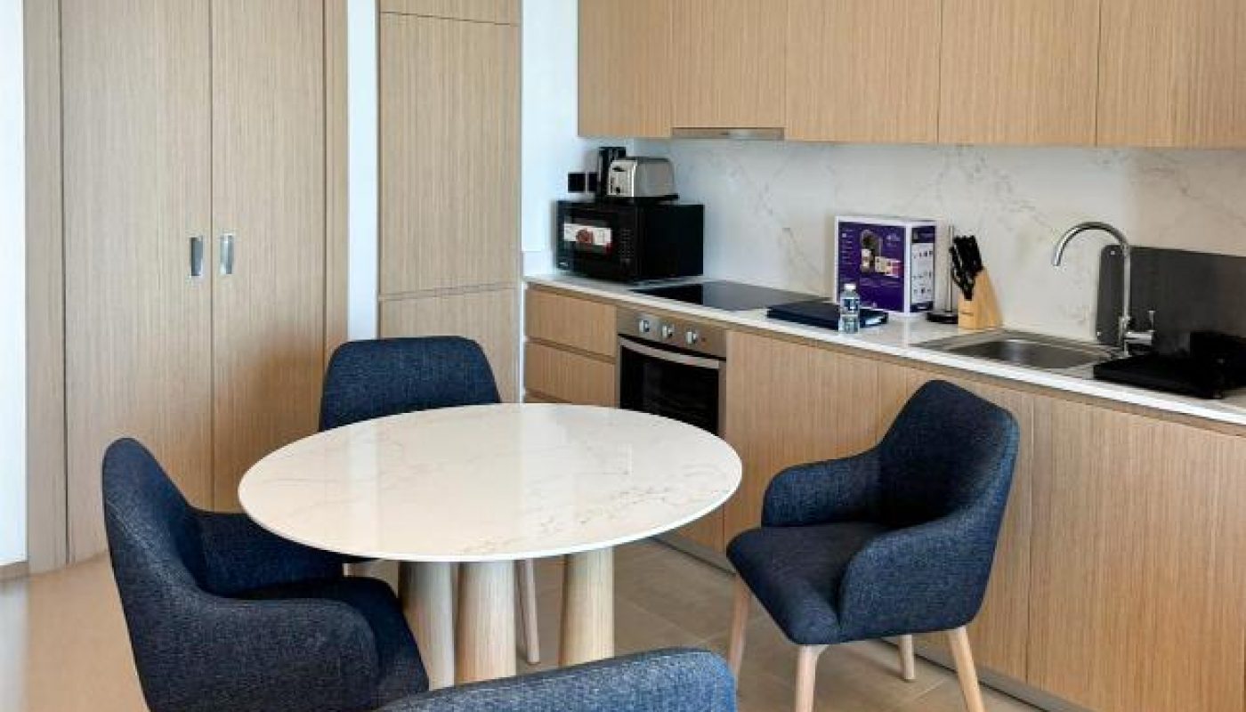 Modern kitchen with light wood cabinets, marble countertop, built-in appliances, and a round table surrounded by four blue upholstered chairs on beige tile flooring.