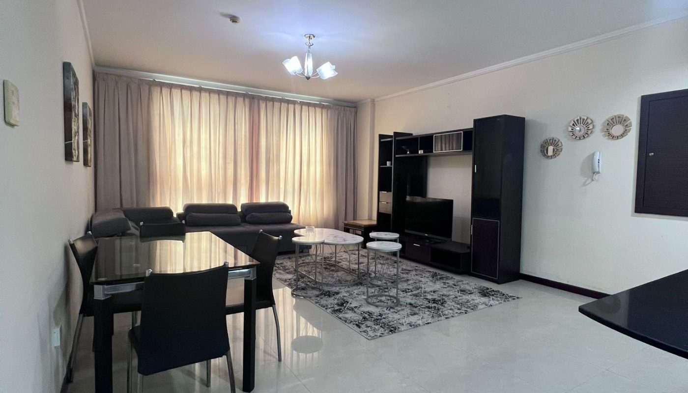 Modern living and dining room with tile flooring, glass dining table, black chairs, dark TV unit, and a large window with beige curtains letting in natural light.