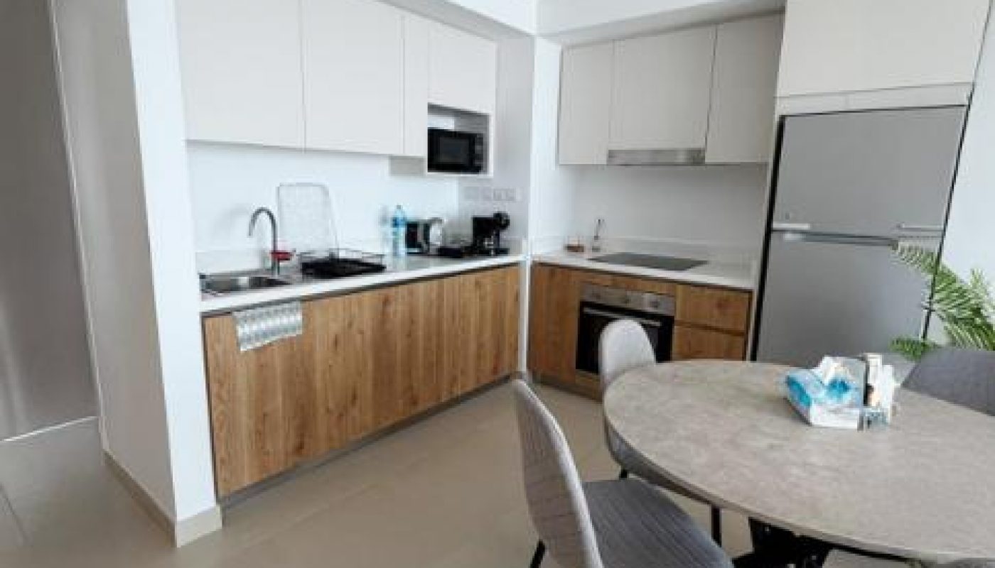 Modern kitchen with wood and white cabinets, built-in appliances, round dining table with four grey chairs, and a refrigerator; various utensils and supplies are on the countertop and table.