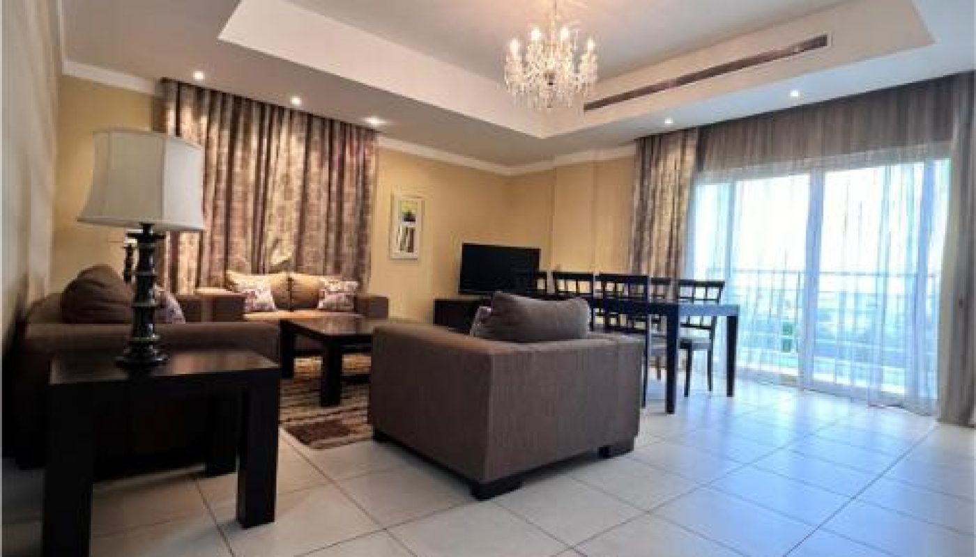 Spacious living and dining area with brown sofas, a dining table, a lamp, a TV, and large windows with sheer curtains letting in natural light.