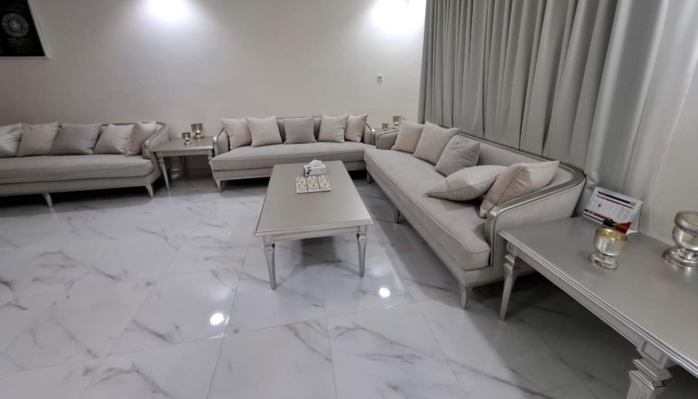 Modern living room with white marble floor, light gray sofas, matching tables, decorative pillows, and neutral drapes. Minimalist decor with soft, ambient lighting.