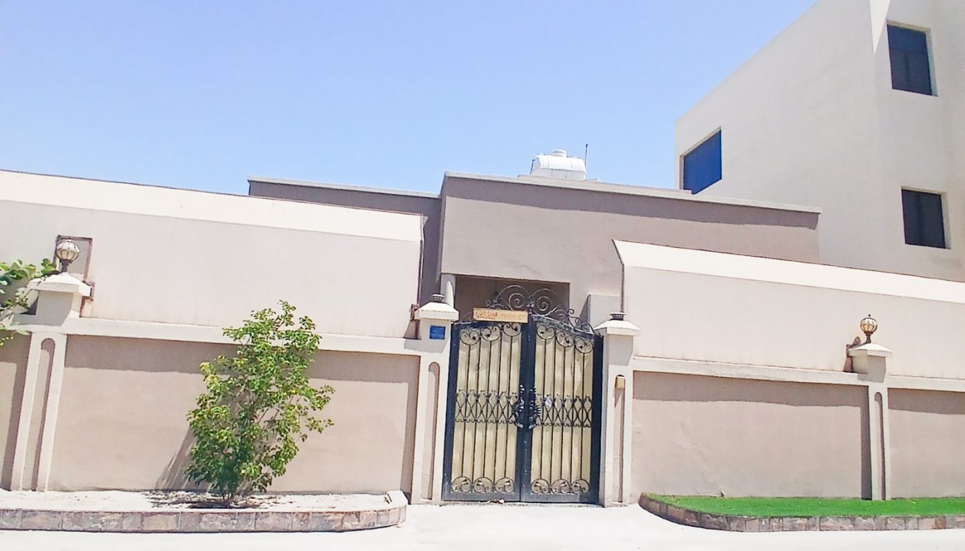 A modern beige building with a high wall, black ornamental gate, small shrubs, and a clear blue sky in the background.