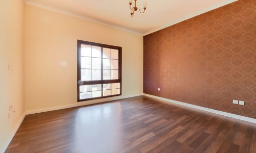 Empty room with wooden floor, textured wallpaper accent wall, large window with a grid pattern, and a small, simple chandelier in a 3BR villa located in Al Areen with access to a big garden.