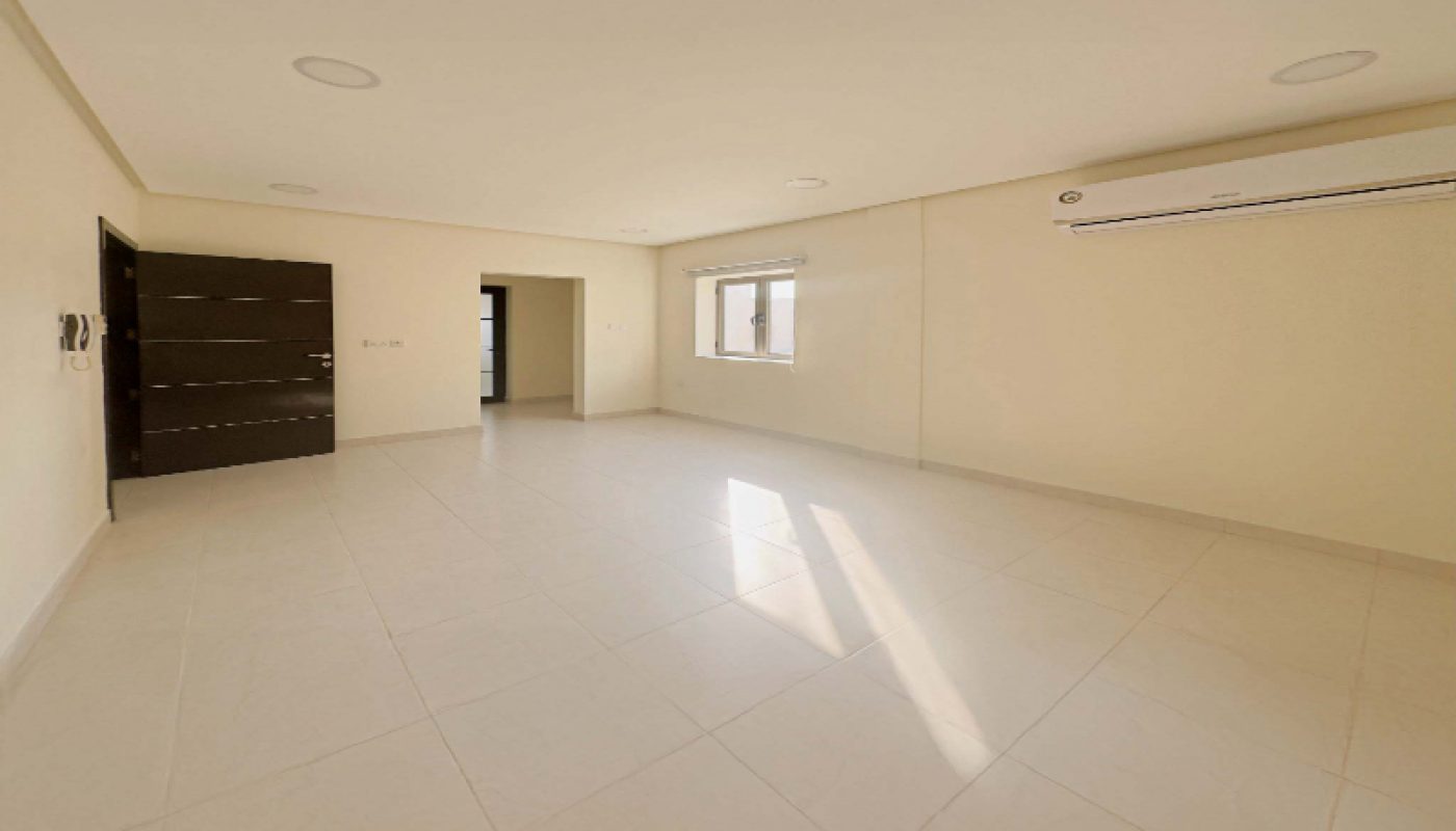 Empty, unfurnished room with white tiled flooring, cream-colored walls, a dark door, a window letting in sunlight, and a wall-mounted air conditioner.