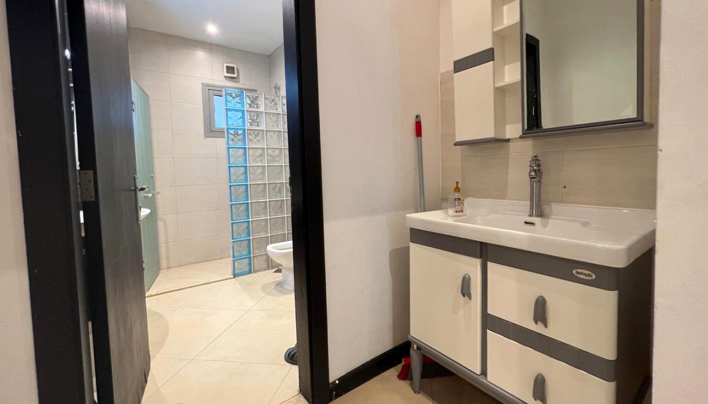 Bathroom with a white vanity, mirror cabinet, and mop; door open to reveal toilet and glass block partition in adjoining room.