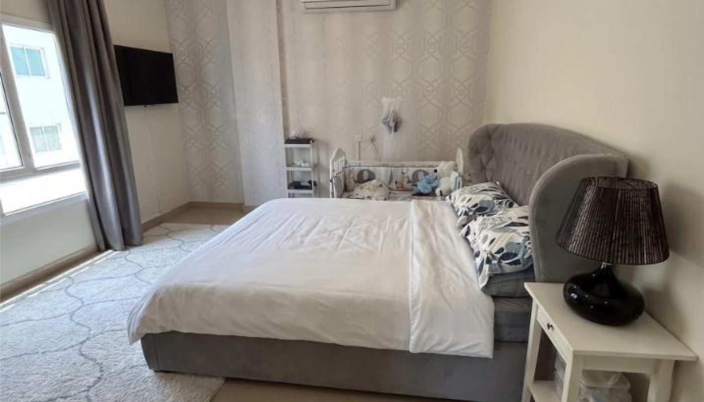A modern bedroom with a large bed, white bedding, bedside table with a black lamp, wall-mounted TV, window with curtains, and an air conditioner above the bed.