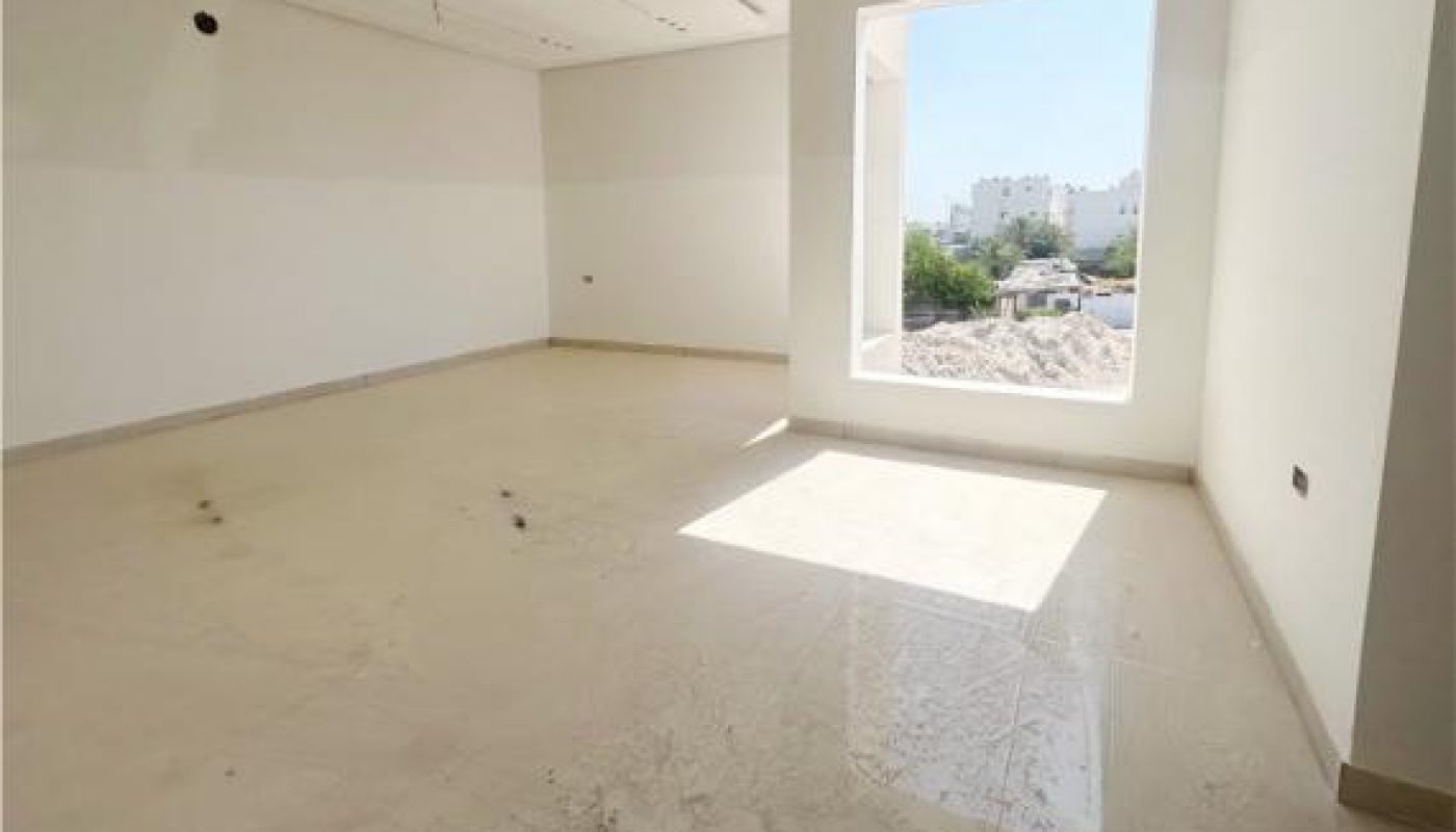 Empty, unfurnished room with off-white walls and a large window letting in natural light; the floor appears to have some dirt and debris.