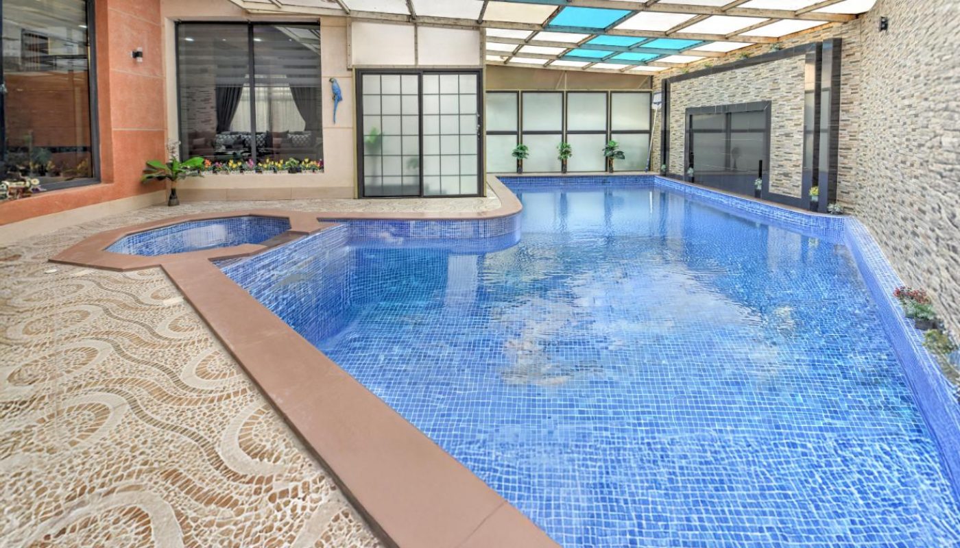 Indoor swimming pool with blue tiles, attached hot tub, stone-patterned flooring, glass walls, and a partially transparent ceiling.