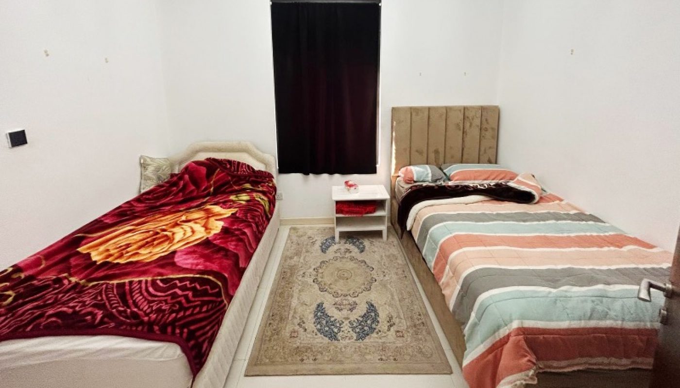 Two single beds with different bedding, a small rug, a chandelier, a covered window, and a table with some items in a simple, white-walled room.