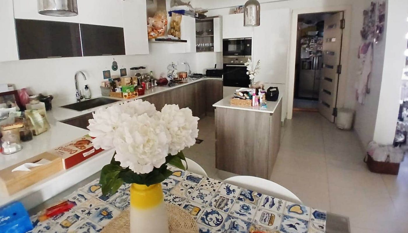 A modern kitchen features white cabinets, a wooden island with a floral tablecloth, and a vase of white flowers. Pendant lights hang overhead, and appliances are visible in the background.