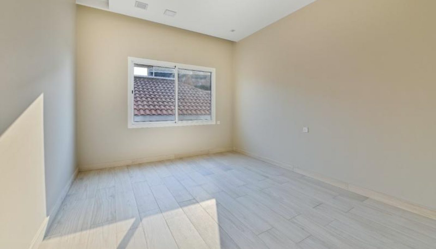 Empty room with beige walls, light-colored wooden flooring, a white ceiling, and a window overlooking a tiled roof and some outdoor structures.