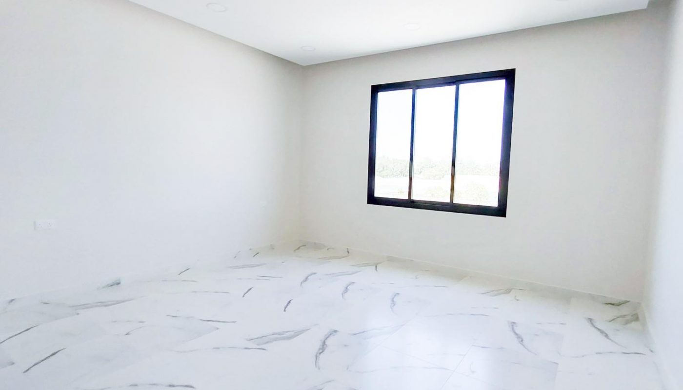 Empty room with white walls, a large window, and white marble-patterned floor tiles. Ceiling lights are recessed, and natural light enters through the window.