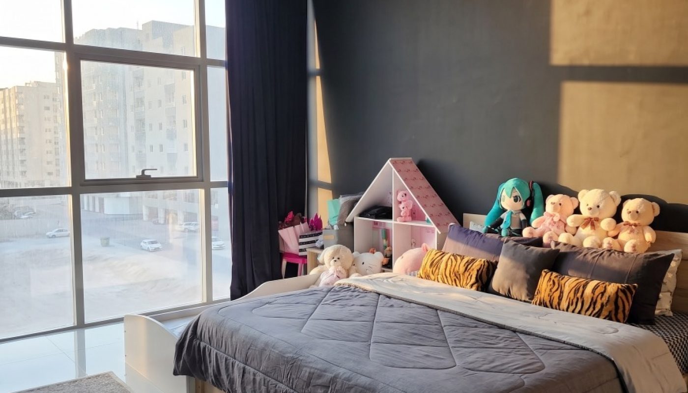 Bedroom with a large window, sunlight streaming in, a bed with plush toys, a dark accent wall, a rug, and a small pink desk in the corner.