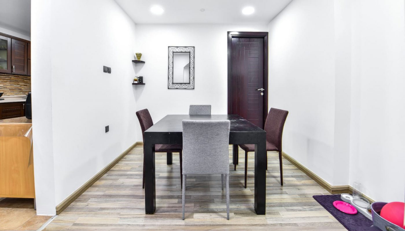 Modern dining room with a black table, four chairs, and a mirror on a white wall. Wooden flooring and a doorway in the background. Gym equipment on the right.