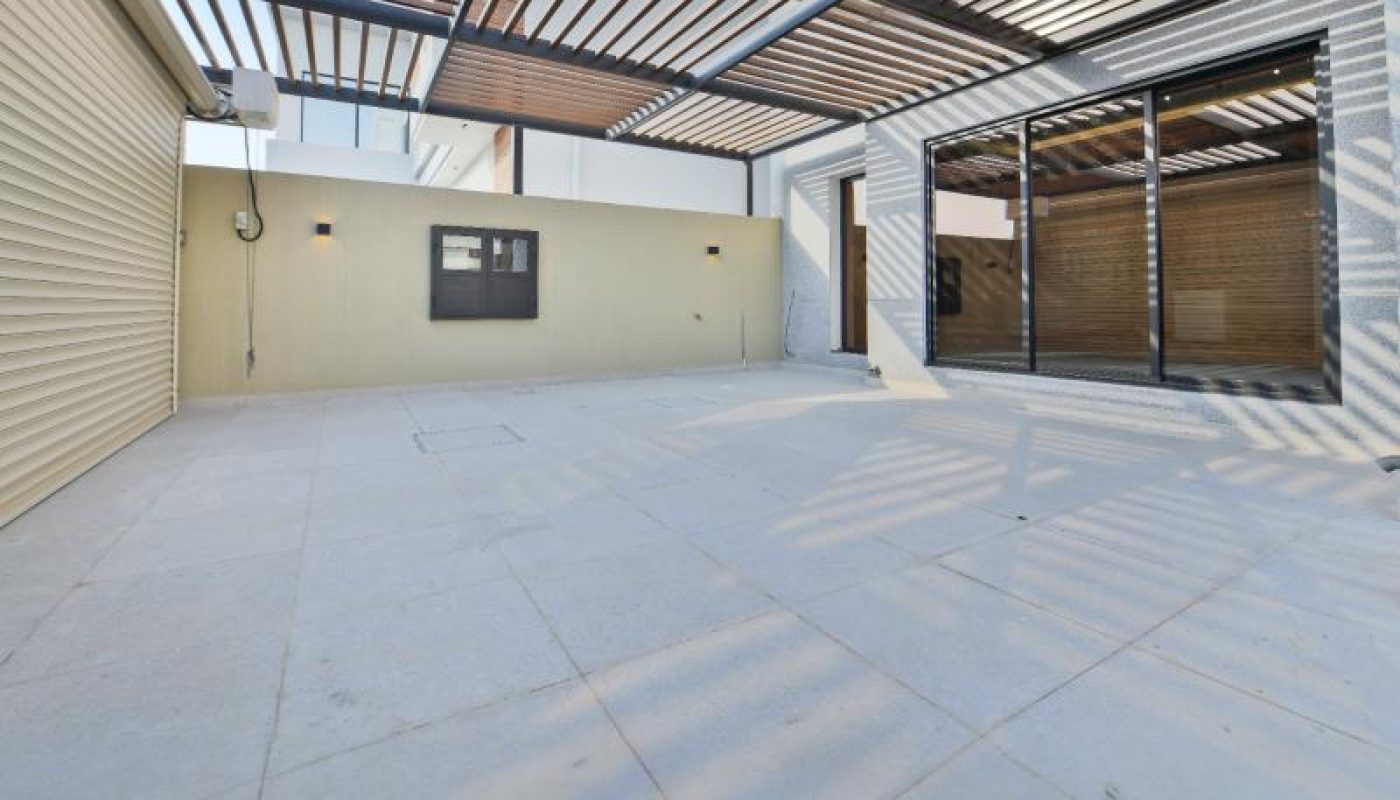 Spacious outdoor patio area with light-colored tiled floor, overhead slatted pergola, and sliding glass doors leading inside.