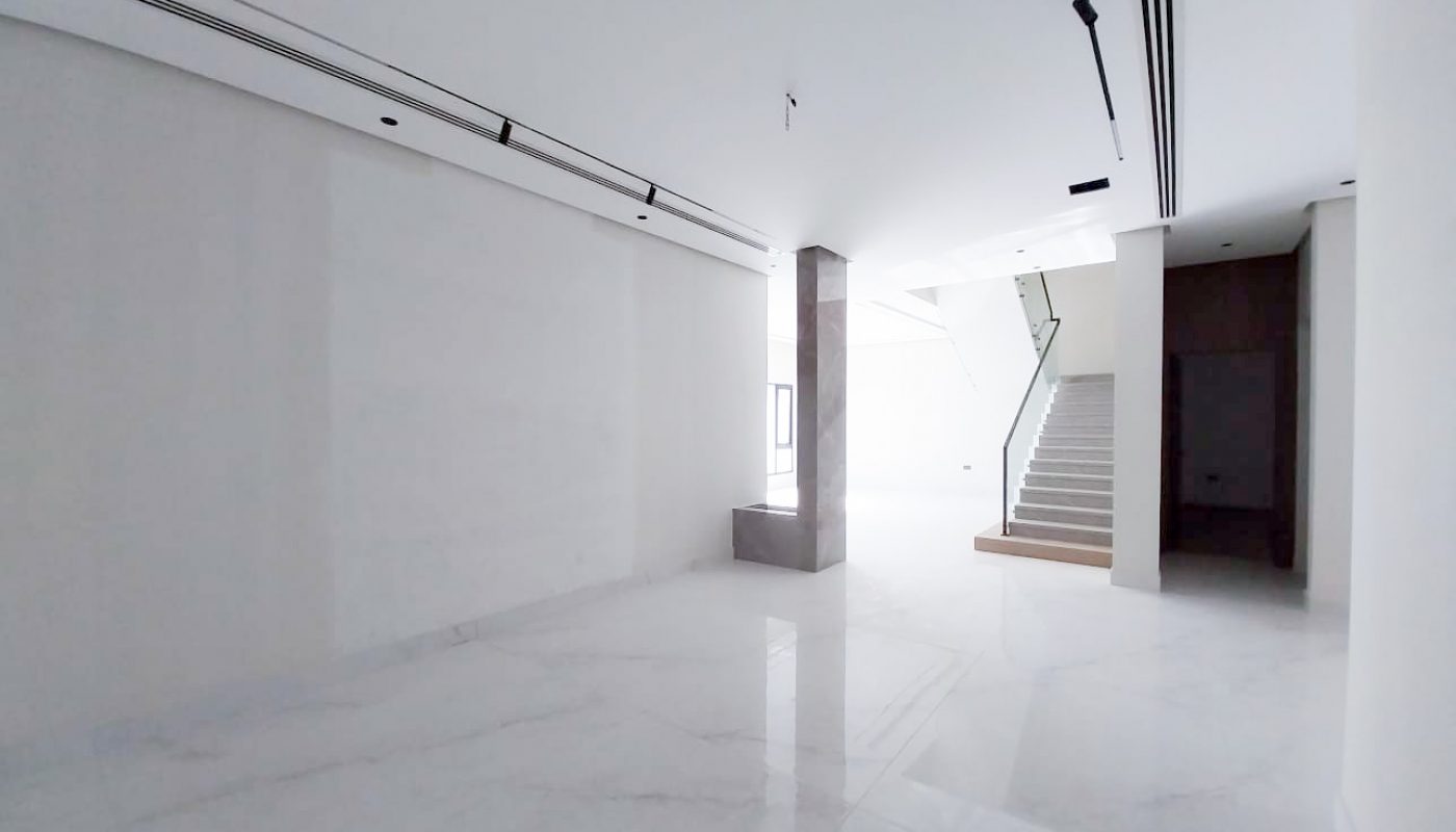 Spacious, empty interior with white marble floors, white walls, a central pillar, large window, and a staircase leading to the upper floor.