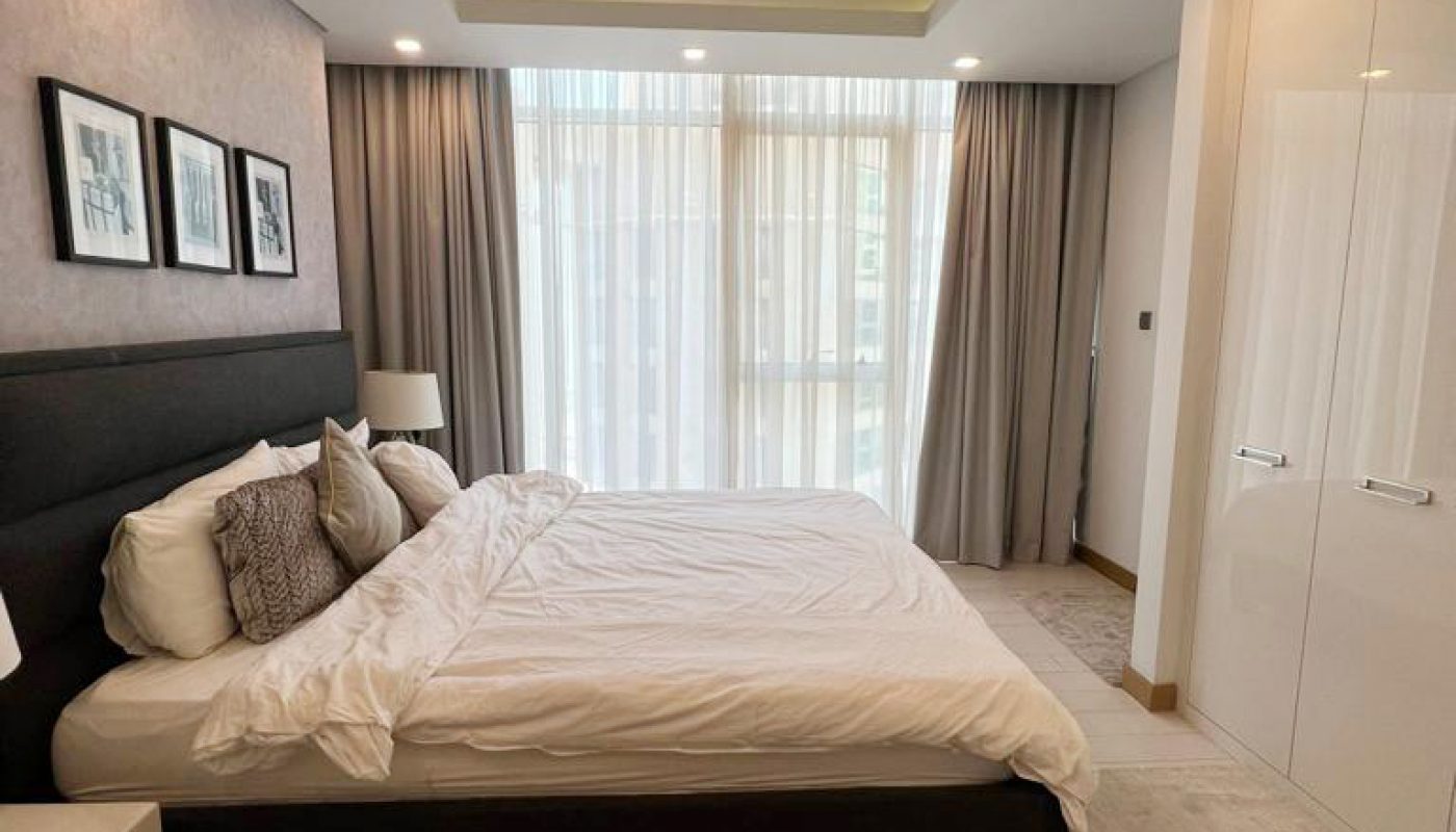 A tidy bedroom with a double bed, bedside tables, wall art above the headboard, floor-to-ceiling window with curtains, and built-in white wardrobe.