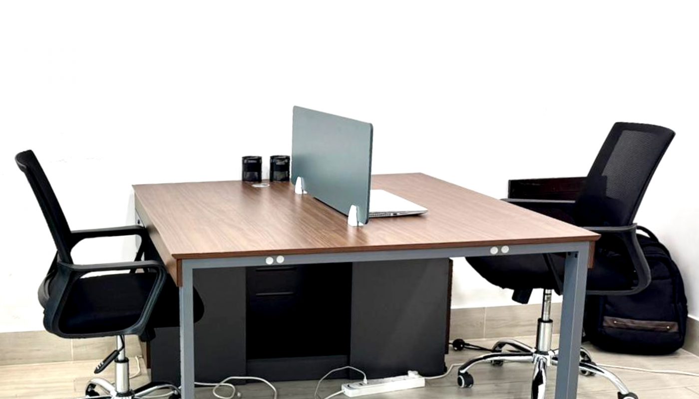 Two black office chairs face each other across a wooden desk with a monitor, keyboard, mouse, and mugs. Cables are visible under the desk on a light wood floor.