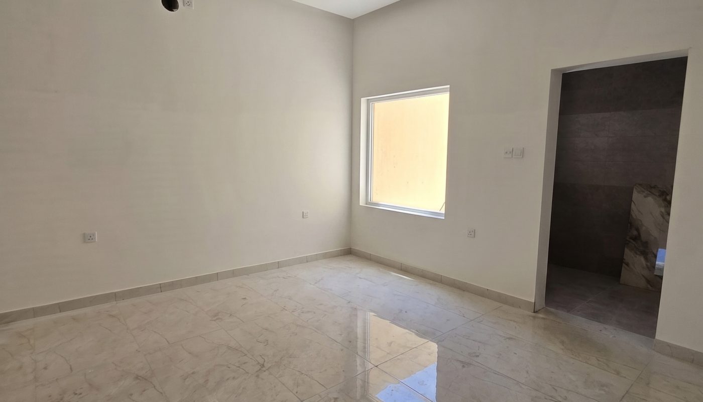 An empty, unfurnished room with tiled floors, white walls, a single window, and an open doorway leading to another space.