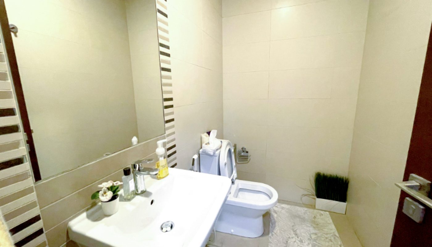 A small bathroom with a wall-mounted sink, a mirror, a toilet, beige tiled walls, and a decorative mat and plant on the floor.
