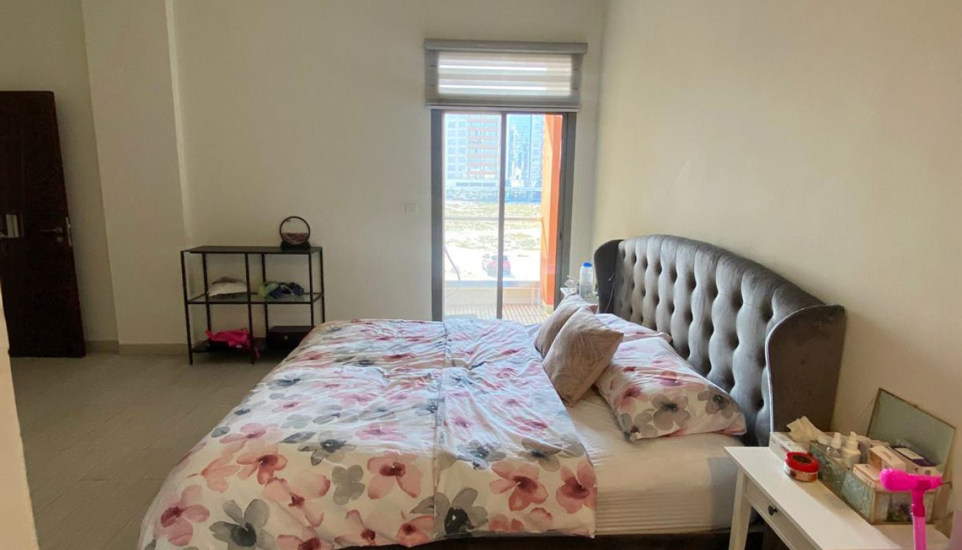 A bedroom with a double bed, floral bedding, a bedside table with items, an open shelf, and a window letting in natural light.