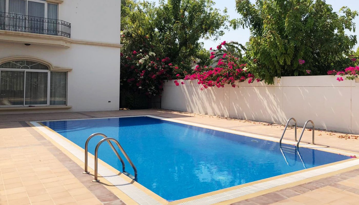A rectangular swimming pool with metal ladders is surrounded by a tiled patio. A spacious 4BR villa for rent in Hamala, featuring a white building with a balcony and large windows on the left side, completes this picturesque scene, with trees and flowers in the background.