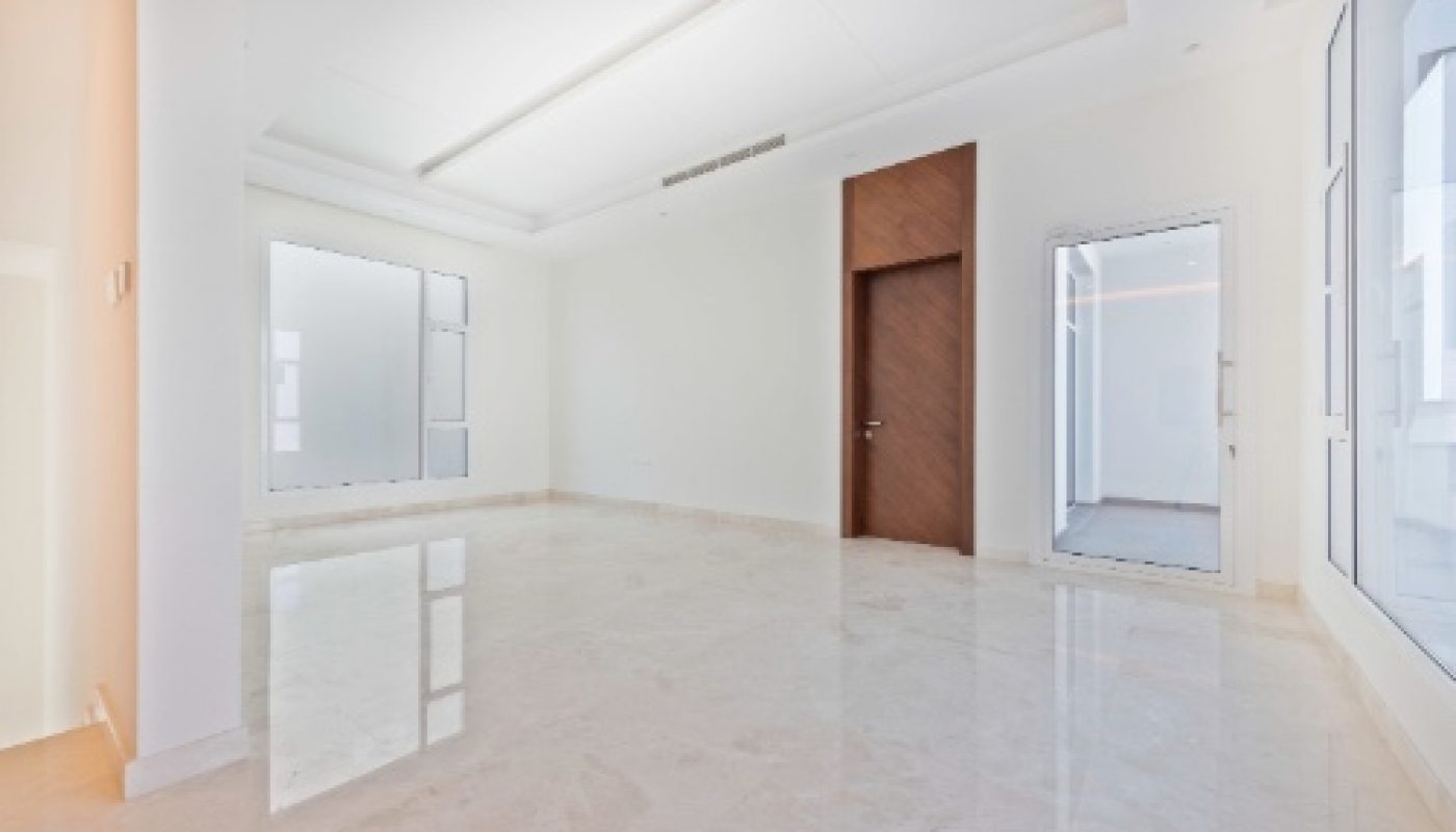 Spacious empty room with white walls, large windows, marble floor, and a wooden door. Natural light fills the minimalist, modern interior.