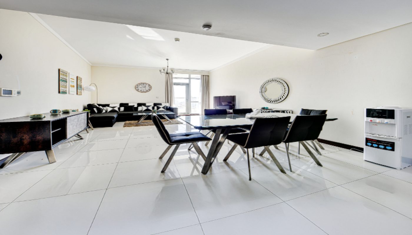 Modern open-plan living and dining area with black chairs, a dining table, white tiled floors, a sofa, wall art, and a water dispenser. Natural light enters through large windows.