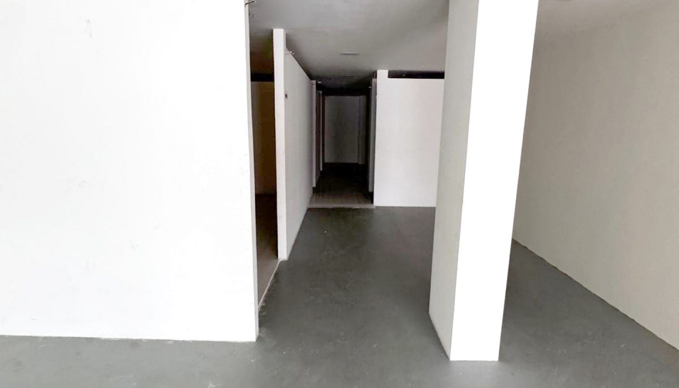 Empty, white-walled interior space with gray concrete floor, columns, and a narrow hallway extending towards the back.