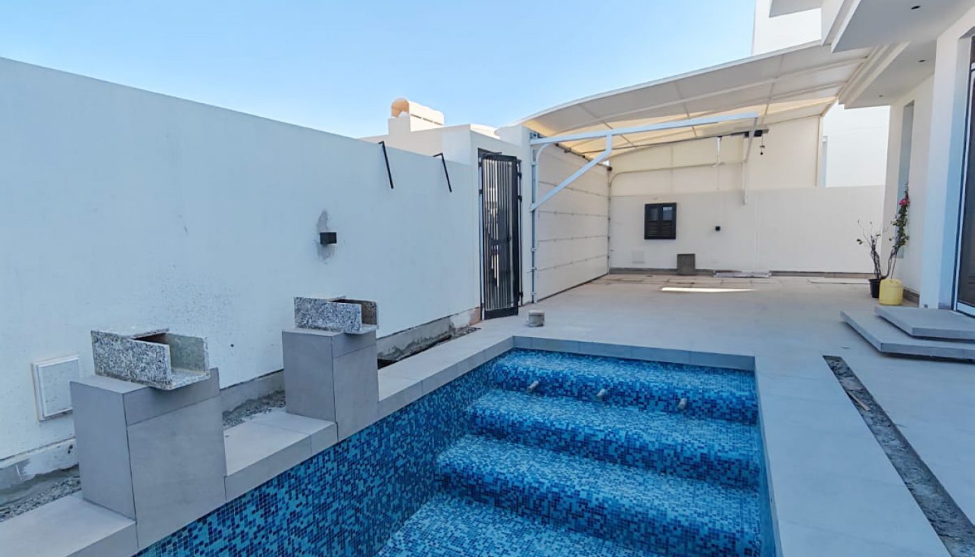 Empty rectangular pool with blue mosaic tiles, built-in steps, and two water features in a modern outdoor patio area under a clear sky.