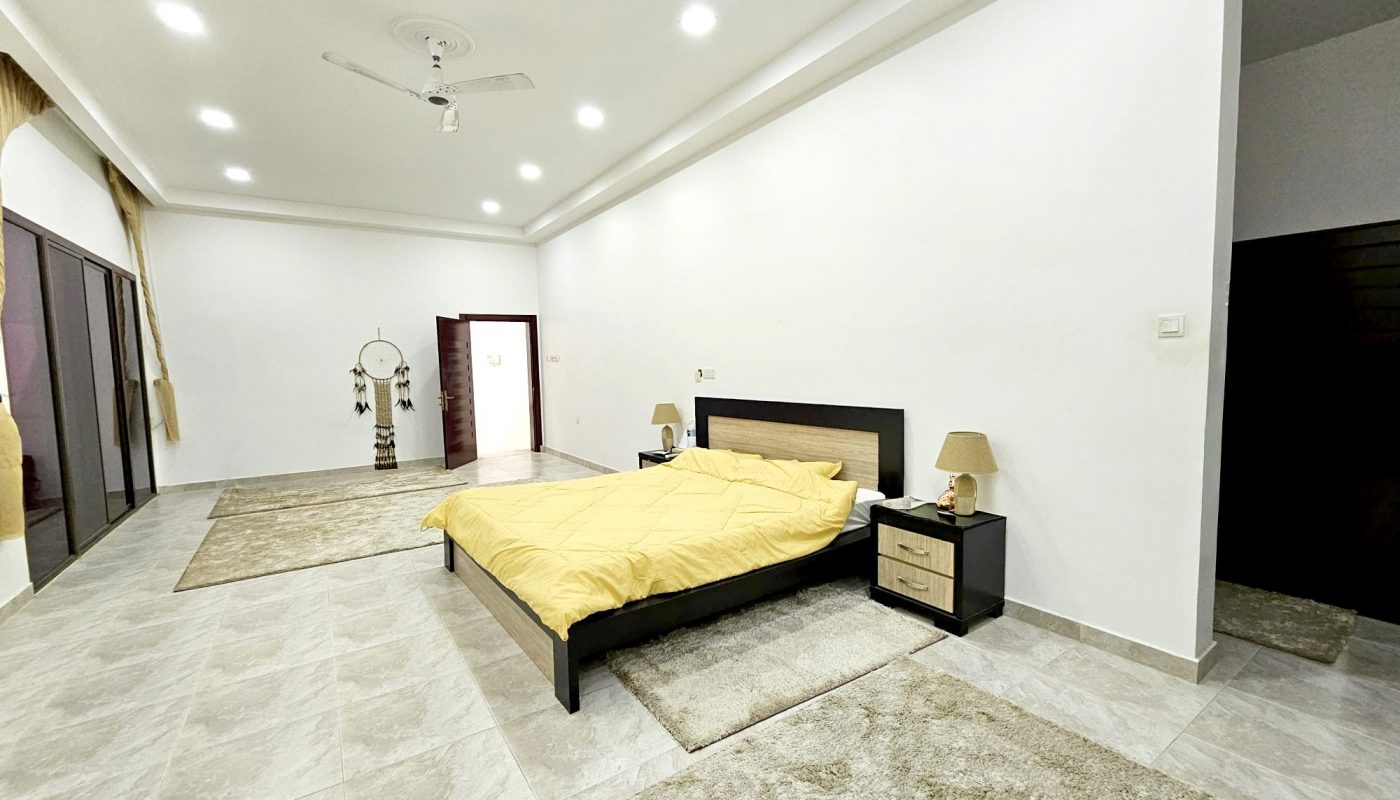 Spacious bedroom with a large bed, two nightstands, lamps, beige rugs, and a ceiling fan, featuring tiled floors and white walls.