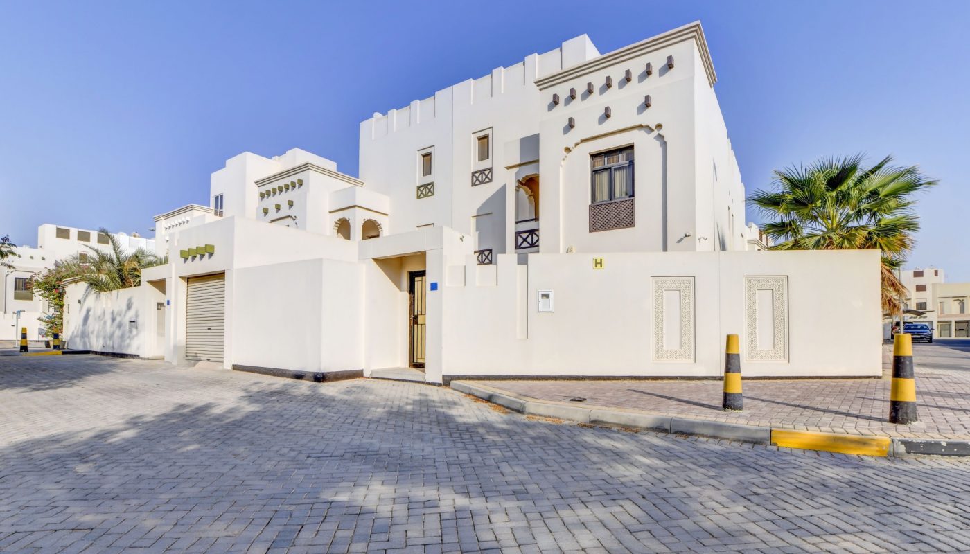 Modern white villa with decorative windows, palm trees, and a paved driveway under a clear blue sky.