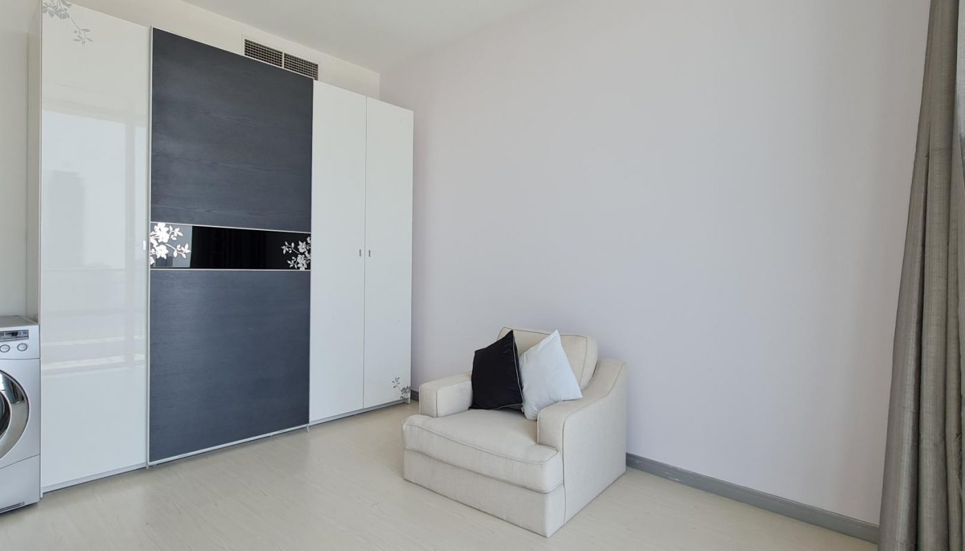 A modern, minimalistic room with a washing machine, a large wardrobe, a beige armchair with cushions, and a window with grey curtains.