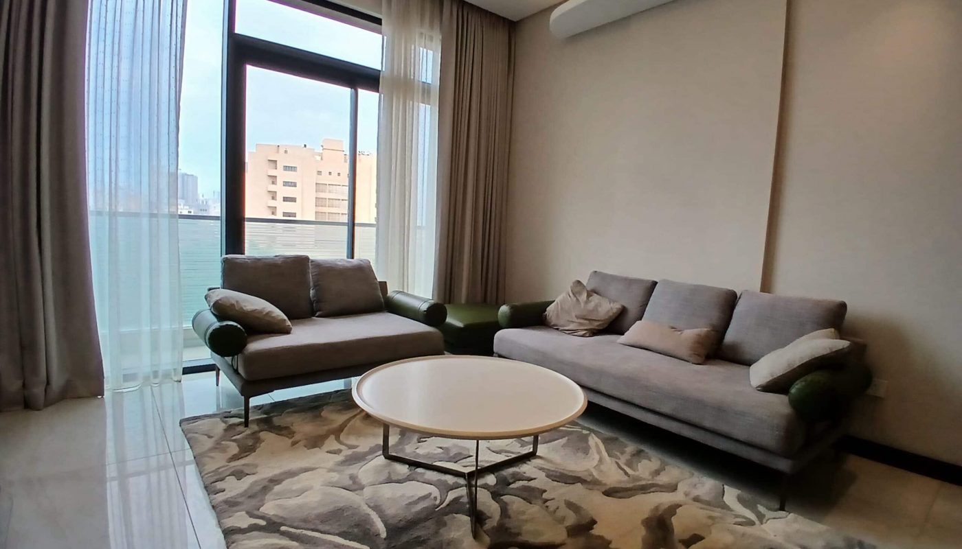 Modern living room interior with a gray sofa, matching armchair, round coffee table, patterned rug, and large window offering a Mahooz city view.