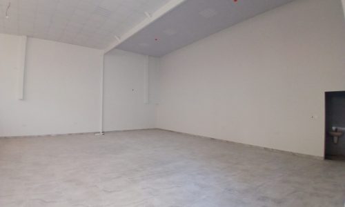 Empty commercial space with gray tiled floor and white walls, showing auto draft ceiling lights and a small door on the right.