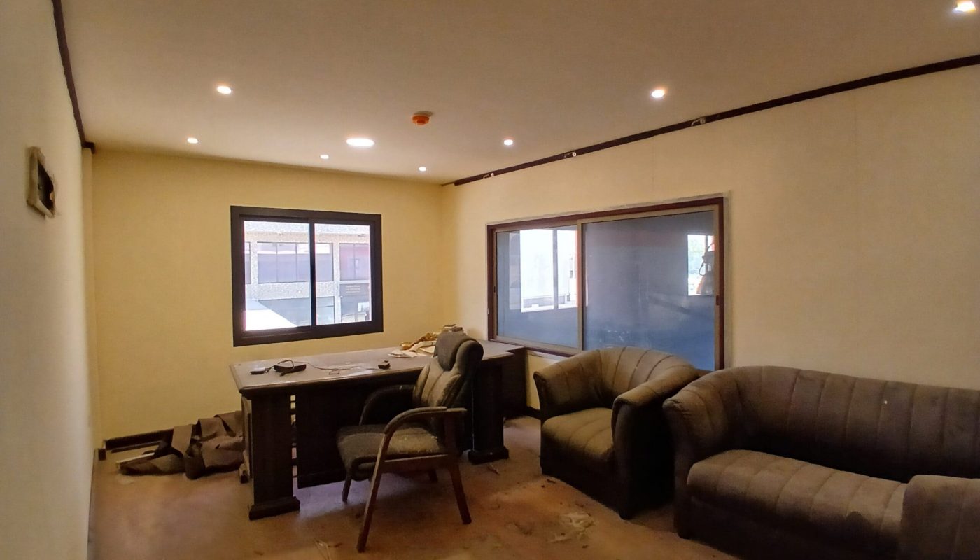 Sparsely furnished semi-fitted shop with two grey sofas, a desk with a chair, scattered debris on the floor, and ceiling spotlights, viewed during daylight.